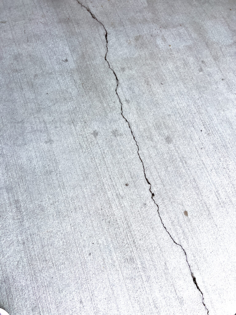 Close-up of a cracked concrete surface, showing distinct lines and fractures. The texture of the concrete is clearly visible, highlighting the wear and damage over time. This image captures the raw and rugged nature of deteriorating concrete, often seen in sidewalks, driveways, and other paved areas.
