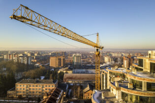 Apartment or office tall building under construction, top view. Tower crane on bright blue sky copy space background, city landscape stretching to horizon. Drone aerial photography.