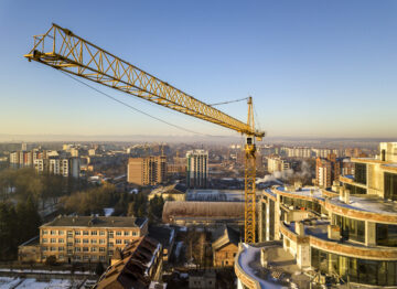Apartment or office tall building under construction, top view. Tower crane on bright blue sky copy space background, city landscape stretching to horizon. Drone aerial photography.