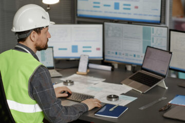 Back view of male construction engineer wearing hardhat at workplace in office and using computer, copy space