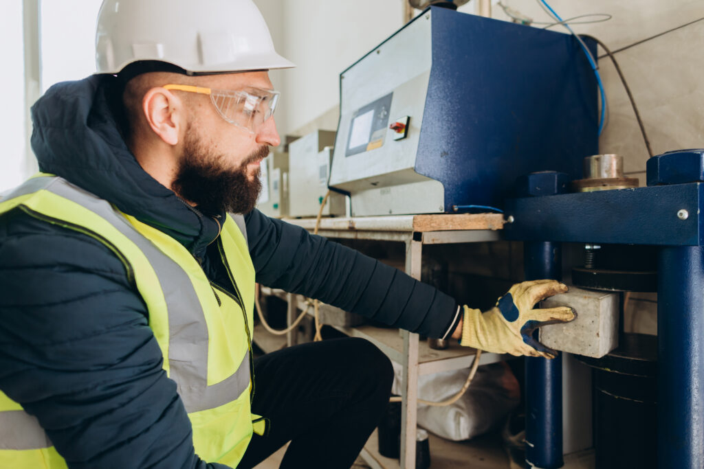Quality control engineer placing concrete cube into compression testing machine in factory laboratory.